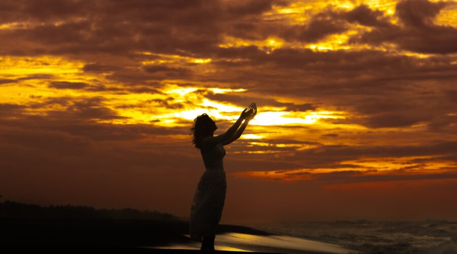 silhouette of a woman standing on the beach with her arms raised at sunset